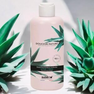 Un flacon de Shampooing Douceur Sublimo Aloé Vera 250 ml est présenté. Ce shampooing sublimateur de boucles est élaboré à partir de 96 % d'ingrédients naturels, dont l'aloe vera. Le flacon présente un motif de feuilles d'aloe vera, un bouchon blanc et est posé sur un fond de feuilles d'aloe vera supplémentaires.