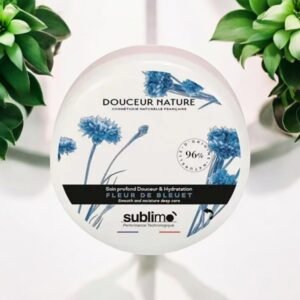 Un contenant rond étiqueté « Soin Sublimo Soin Profond Douceur & Hydratation Fleur de Bleuet 150 ml » présente des fleurs bleues sur le devant, plus précisément « Fleur de Bleuet ». Il est indiqué « 96 % d'origine naturelle », « Soin profond lissant et hydratant » et « Sublimo Performance Technologique », avec des plantes vertes en arrière-plan.