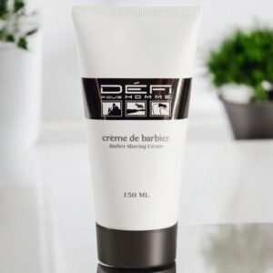 Un tube de crème à raser « Défi pour Homme » est posé sur une surface réfléchissante. Le tube est blanc avec une étiquette noire et argentée, comportant une iconographie d'articles liés au rasage. La capacité est de 150 ml. Des plantes floues sont visibles en arrière-plan.