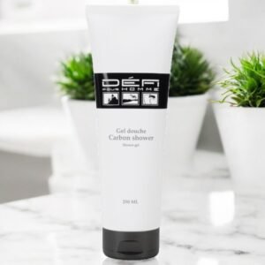 Un tube de gel douche Défi Pour Homme Carbon est posé sur une surface en marbre. Le tube blanc au bouchon noir porte bien en évidence le logo et le texte de la marque. En arrière-plan, des plantes vertes ajoutent une touche de nature à ce décor épuré et moderne.