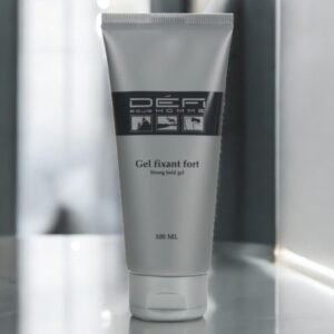 Un tube argenté de 100 ml de gel fixant fort Défi pour Homme est présenté debout sur une surface réfléchissante sur un fond flou. L'étiquette comporte des icônes et du texte en noir et blanc.