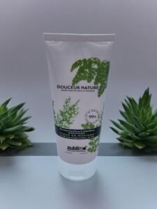 Un tube blanc de « BB crème capillaire Sublimo Fleur de Moringa 100 ml » avec une image de feuilles vertes sur l'étiquette. La crème est enrichie en fleur de moringa et prétend être d'origine naturelle à 99 %. Le tube est placé entre deux petites plantes succulentes sur une surface blanche sur fond gris.