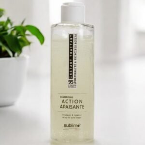 Une bouteille transparente de shampoing Sublimo Professionnel étiquetée « Action Apaisante » se trouve sur une surface blanche. La bouteille contient un liquide jaune pâle et possède un bouchon blanc. En arrière-plan, on voit une plante verte légèrement floue.