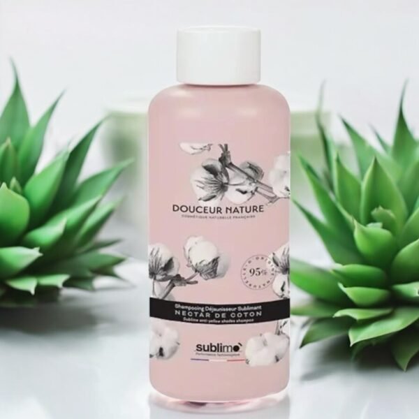 Une bouteille de Shampoing Douceur Sublimo Nectar de Coton 250 ml est centrée sur l'image. La bouteille est rose avec un motif floral et un texte de marque sur le devant. Deux plantes succulentes vertes sont placées de chaque côté de la bouteille en arrière-plan, ajoutant une touche de nature à la scène.