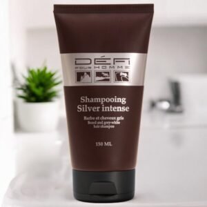 Au premier plan, un flacon de 150 ml de Défi Pour Homme Shampooing Silver Intense pour barbe et cheveux gris-blancs est exposé. Le flacon marron est orné d'une étiquette argentée avec du texte et des icônes. Une plante floue et des accessoires de salle de bains sont visibles à l'arrière-plan.