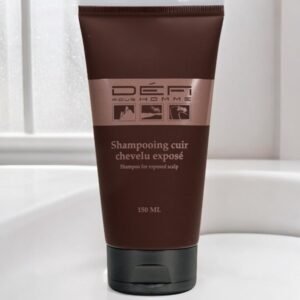 Un tube brun de 150 ml de shampoing pour cuir chevelu exposé Défi Pour Homme est posé à la verticale sur une surface blanche sur fond de salle de bain flou. L'étiquette comporte du texte en français et en anglais.