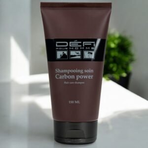 Un tube de 150 ml de shampooing capillaire Carbon Power de Defi Pour Homme est présenté. Le tube est marron avec une étiquette noire comportant le nom de la marque, la description du produit et trois icônes des étapes de soins capillaires. Une plante verte est visible sur l'arrière-plan flou.