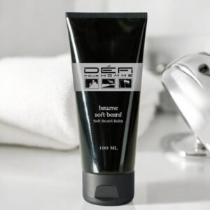 Un tube noir de baume à barbe doux Défi Pour Homme (100 ml) est mis en valeur. Le tube comporte du texte et de petites illustrations d'icônes. Une serviette blanche et une partie d'un accessoire de salle de bains chromé sont floues en arrière-plan.