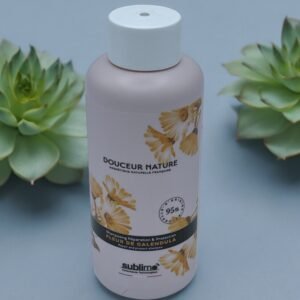 Une bouteille beige étiquetée « Découvrez les bienfaits du shampoing Douceur Sublimo Fleur de Calendula » avec un motif floral se dresse entre deux plantes succulentes vertes. L'étiquette mentionne « 95 % » et « Fleur de Calendula », à côté de la marque « Sublimo » en bas. Le fond est d'un bleu doux et discret.