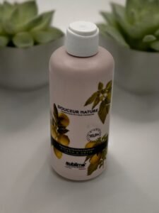 Une bouteille de Shampoing Sublimo Citron et Orties 250 ml est posée sur une surface, avec un bouchon blanc et une étiquette représentant des agrumes et des feuilles. L'étiquette indique que le shampoing est parfumé aux agrumes et composé à 93,8 % d'ingrédients naturels, avec deux plantes succulentes en arrière-plan.