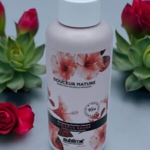 Une bouteille de Shampooing Sublimo Hibiscus Rouge 250 ml avec un motif floral sur l'étiquette est posée sur une surface grise. Autour de la bouteille se trouvent des roses rouges et des plantes succulentes vertes, ajoutant une ambiance naturelle et fraîche au décor. La bouteille prétend contenir 95 % d'ingrédients naturels.