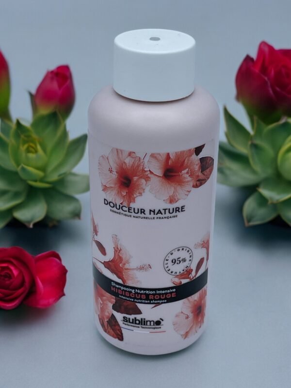 Une bouteille de Shampooing Sublimo Hibiscus Rouge 250 ml avec un motif floral sur l'étiquette est posée sur une surface grise. Autour de la bouteille se trouvent des roses rouges et des plantes succulentes vertes, ajoutant une ambiance naturelle et fraîche au décor. La bouteille prétend contenir 95 % d'ingrédients naturels.