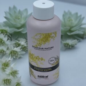 Un flacon rose de "Shampooing Sublimo Fleur de Mimosas 250ml" est présenté sur un fond blanc doux entouré de plantes succulentes. Le flacon présente des motifs floraux jaunes et se revendique comme étant naturel à 95 %.