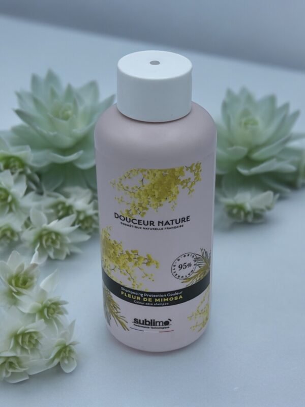 Un flacon rose de "Shampooing Sublimo Fleur de Mimosas 250ml" est présenté sur un fond blanc doux entouré de plantes succulentes. Le flacon présente des motifs floraux jaunes et se revendique comme étant naturel à 95 %.