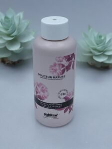 Un flacon rose clair de shampoing « Shampooing Sublimo Rose de Damas 250 ml » est présenté avec des accents de motifs floraux et une étiquette indiquant 93 % d'ingrédients naturels. Le flacon repose sur une surface grise avec deux plantes succulentes en arrière-plan, ajoutant une touche de verdure.