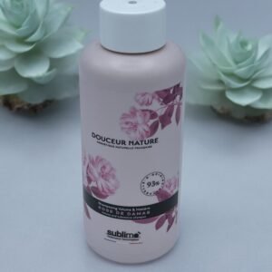 Un flacon rose clair de shampoing « Shampooing Sublimo Rose de Damas 250 ml » est présenté avec des accents de motifs floraux et une étiquette indiquant 93 % d'ingrédients naturels. Le flacon repose sur une surface grise avec deux plantes succulentes en arrière-plan, ajoutant une touche de verdure.