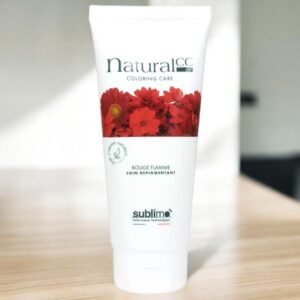 Un tube blanc étiqueté « Natural CC Coloring Care Rouge Flamme » avec des fleurs rouges, posé sur une surface en bois. Le nom de la marque « Sublimo » apparaît au bas du tube. La lumière du soleil illumine la scène depuis l'arrière gauche, projetant une ombre douce.