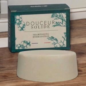 Une boîte rectangulaire étiquetée « Douceur Solide Shampooing Hydratation » avec des illustrations botaniques est placée derrière une barre de shampoing solide blanche en forme de dôme sur une surface en bois. La boîte et la barre de shampoing sont contre un mur blanc.