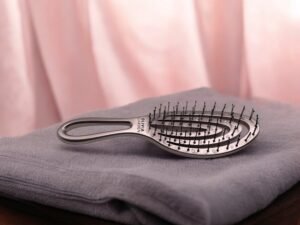 Une brosse à cheveux argentée, semblable à la Brosse Olivia Garden Rose (Copie), avec son design ouvert et ses poils distinctifs, repose sur une serviette grise pliée. Des rideaux rose pâle en arrière-plan créent une atmosphère calme et élégante.