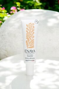 Un tube blanc de Enaya CC Crème Perfection, présenté à la verticale sur une surface extérieure avec un fond vert et floral flou. L'emballage est minimaliste avec un design rappelant une feuille d'or et un texte noir, soulignant sa protection SPF de 30 % et son origine française.