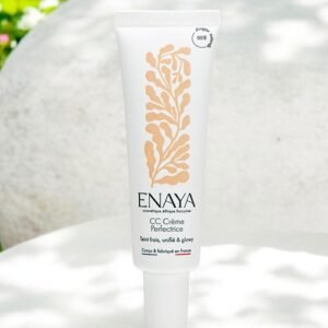 Un tube blanc de Enaya CC Crème Perfection, présenté à la verticale sur une surface extérieure avec un fond vert et floral flou. L'emballage est minimaliste avec un design rappelant une feuille d'or et un texte noir, soulignant sa protection SPF de 30 % et son origine française.