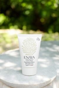 Un tube blanc de crème pour les mains Nourishing Velvet d'Enaya est placé sur une surface en pierre à l'extérieur, avec de la verdure en arrière-plan flou. Le tube présente un dessin botanique simple et élégant et un texte minimaliste détaillant les informations sur le produit.