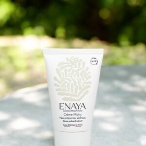 Un tube blanc de crème pour les mains Nourishing Velvet d'Enaya est placé sur une surface en pierre à l'extérieur, avec de la verdure en arrière-plan flou. Le tube présente un dessin botanique simple et élégant et un texte minimaliste détaillant les informations sur le produit.