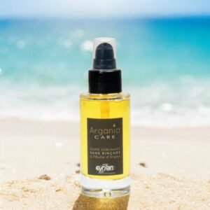 Un flacon d'huile capillaire Argania Care Elixir Sublimant Sans Rinçage se dresse sur une plage de sable avec l'océan et le ciel bleu en arrière-plan. Le flacon est transparent avec une pompe noire et contient de l'huile jaune doré.