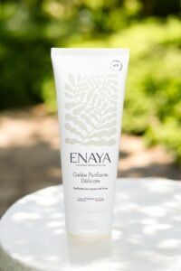 Un tube de Gelée Purifiante Délicate d'Enaya est posé sur une surface blanche avec un fond vert naturel flou. Le tube est principalement blanc, avec un motif botanique subtil et un texte détaillant le nom et les bienfaits du produit.