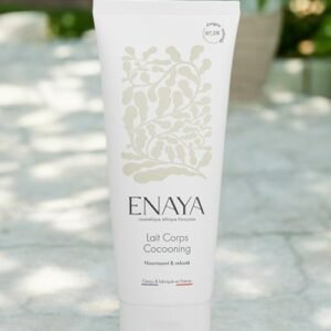 Un tube blanc de lotion pour le corps de la marque Enaya étiqueté « Lait Corps Cocooning » est exposé à l'extérieur sur une surface blanche. L'emballage présente un motif botanique et un texte en français, notamment des phrases telles que « Nourrissant & Velouté » et « Cosmétique Naturel et Biologique ».