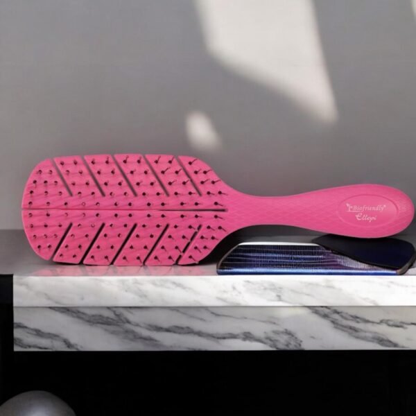 La Brosse Biofriendly-Rose, une brosse à cheveux rose au design texturé et aéré, est posée sur une surface carrelée noire et blanche. Le manche porte le texte « Ecofriendly Edge ».