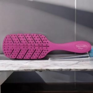 La Brosse Biofriendly-Violet, une brosse à cheveux rose au design en forme de feuille, repose sur un plan de travail en marbre à côté d'un peigne transparent.