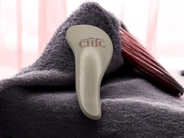 Une brosse à cheveux Brosse Méli-Mélo Chic beige repose sur une serviette douce gris foncé, d'où ressortent ses poils rouges. Le fond est orné de rideaux rose tendre, ajoutant une touche cosy à la scène.