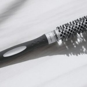 La Brosse Ronde Termix-Diam 17, au design cylindrique avec des poils uniformément répartis autour de son corps en métal et d'un manche noir avec une poignée texturée, repose sur une surface claire. Des ombres sont projetées sur la surface.