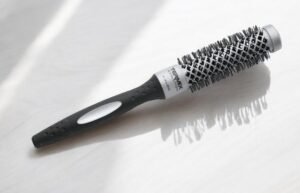 La Brosse ronde Termix-Diam 23, dotée d'un manche noir et d'un corps cylindrique recouvert de poils, repose sur une surface claire. Les ombres projetées par ses poils accentuent le dessin texturé de la brosse.