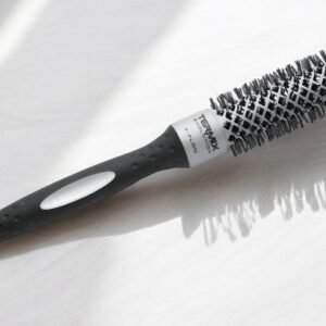 La Brosse ronde Termix-Diam 23, dotée d'un manche noir et d'un corps cylindrique recouvert de poils, repose sur une surface claire. Les ombres projetées par ses poils accentuent le dessin texturé de la brosse.