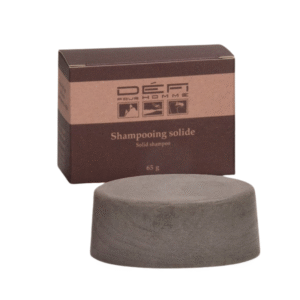 Une boîte marron et beige étiquetée « Shampoing solide-Défi pour Homme - 65 gr » se trouve derrière une barre de shampoing solide ronde et grise pour homme, tous deux présentés sur un fond blanc.