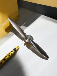 Une Coupe Papier Hélice métallique -DPH, ressemblant à une pale de ventilateur, repose sur une surface blanche, projetant une ombre. Un stylo à points jaunes et noirs se trouve à proximité. Un objet jaune est partiellement visible à l'arrière-plan.