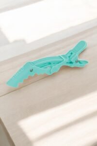 Une pince à cheveux Pince de séparation-Gator X3 verte-Sibel bleu sarcelle, présentant un design en zigzag, est ouverte sur une surface en bois clair, projetant des ombres douces.