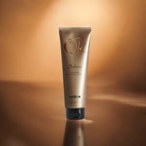 Un tube doré étiqueté « Masque Restructure et Protège -Solaire-Or Infinite-150 ml » se dresse debout sur une surface brune réfléchissante, sur fond de dégradé chaud et lumineux.