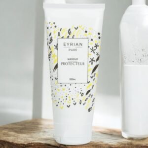 Un tube blanc de Eyrain Pure Masque Protecteur, avec une étiquette de 200 ml, est posé sur une surface en bois. L'étiquette présente des gribouillages décoratifs jaunes, noirs et gris. Une bouteille blanche partiellement visible est visible à l'arrière-plan. Le décor semble minimaliste et bien éclairé.