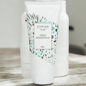 Un tube cylindrique étiqueté « Eyriàn Pure Masque Réparateur » d'un volume de 200 ml est présenté sur une surface en bois. Le tube blanc présente un design abstrait avec des accents bleu sarcelle et marron. Des bouteilles blanches floues sont visibles en arrière-plan.