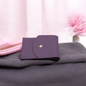 Un petit portefeuille violet repose sur un tissu gris foncé à côté d'une coque de téléphone rose clair. A proximité, le Set onglerie Vert amande (Copie) apporte une touche élégante. En arrière-plan, un pompon rose orne un objet blanc dans un décor aux tons pastels légèrement flous.