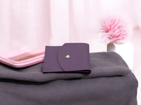 Un petit portefeuille violet repose sur un tissu gris foncé à côté d'une coque de téléphone rose clair. A proximité, le Set onglerie Vert amande (Copie) apporte une touche élégante. En arrière-plan, un pompon rose orne un objet blanc dans un décor aux tons pastels légèrement flous.