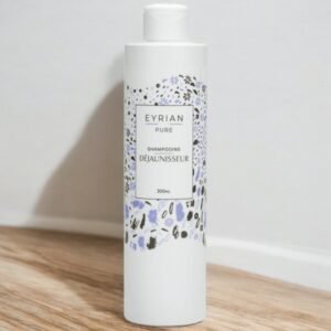 Une bouteille blanche de Eyrien Pure Shampooing Déjaunisseur (300 ml) est posée à la verticale sur une surface en bois. L'étiquette présente de délicates illustrations florales bleues, noires et violettes autour du nom du produit.