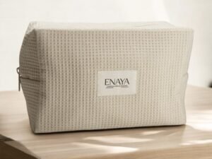 Une trousse de toilette beige Enaya, au design texturé et dotée d'une fermeture éclair sur le côté gauche, repose sur une surface en bois. Le sac est orné d'une étiquette affichant « ENAYA ». Un éclairage doux crée des ombres douces autour de lui.