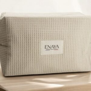 Une trousse de toilette beige Enaya, au design texturé et dotée d'une fermeture éclair sur le côté gauche, repose sur une surface en bois. Le sac est orné d'une étiquette affichant « ENAYA ». Un éclairage doux crée des ombres douces autour de lui.