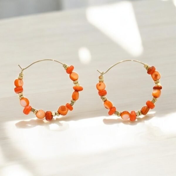 Une paire de Boucles d'oreille créoles nacre / Orange hoop avec de petites perles rondes orange et des accents dorés, élégamment présentées sur une surface de couleur claire avec une douce lumière du soleil projetant des ombres douces.