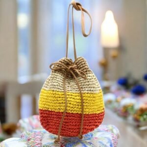 Un petit sac en crochet coloré avec un cordon de serrage est posé sur une table. Ce sac bourse raphia papier tressé présente des rayures horizontales marron, jaune et rouge. Une bougie allumée et un décor flou sont visibles en arrière-plan, créant une ambiance chaleureuse.