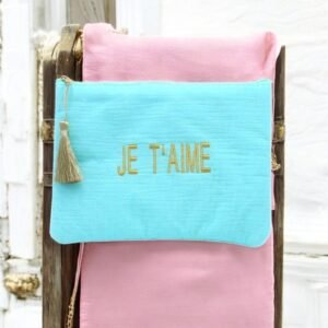 Une trousse à maquillage turquoise « Je t'aime » en gaze de coton, ornée de broderies dorées, est suspendue à une chaise recouverte de tissu rose. Un pompon doré orne la fermeture éclair, contrastant avec un fond de lambris en bois blanc.